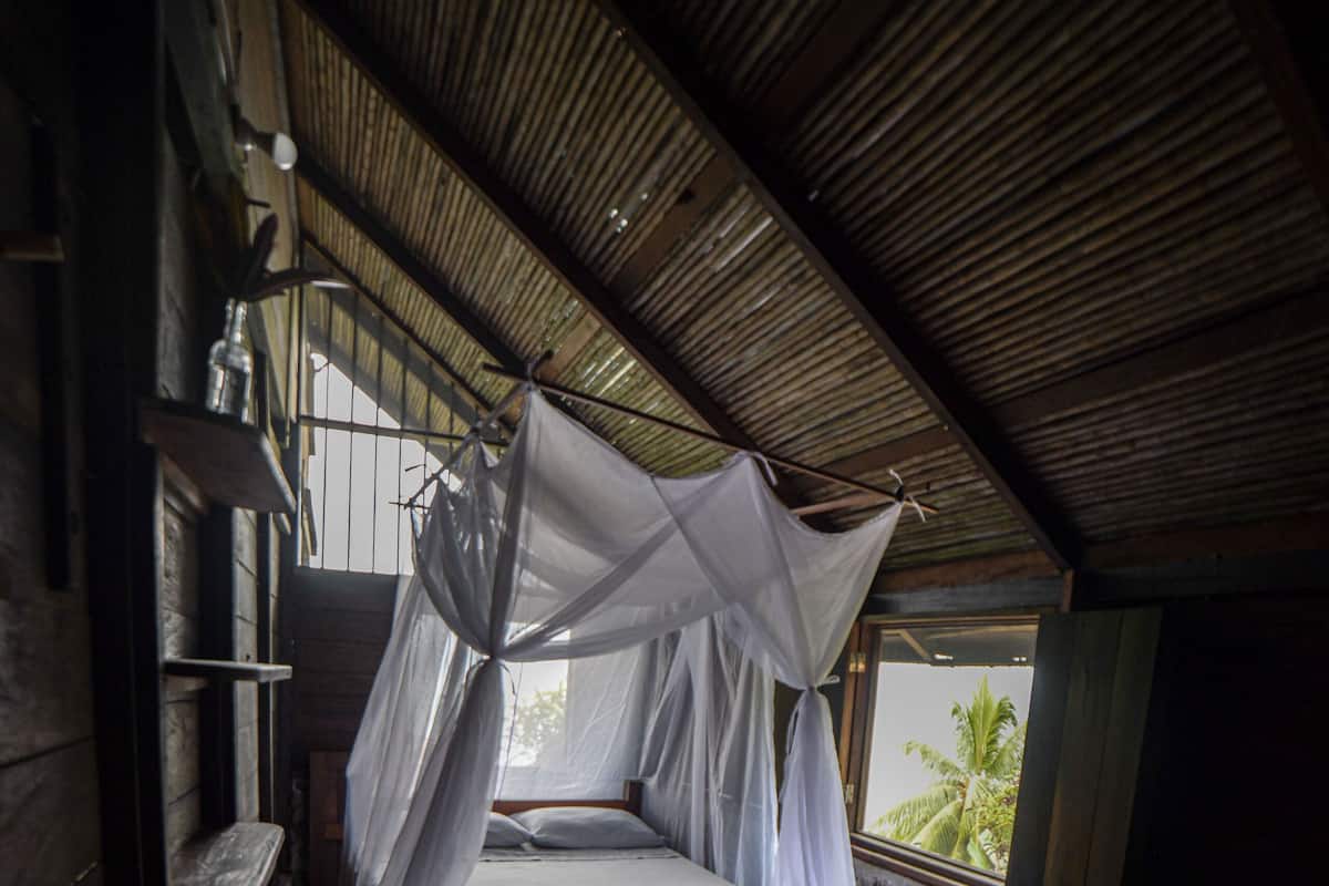 A room with a bed under a mosquito net, a stone wall, and a window with a view of palm trees.