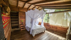Ein Zimmer mit Holzboden, einem Bett unter einem weißen Baldachin, einem kleinen Tisch mit einer Pflanze und einer Treppe zu einer erhöhten Ebene.