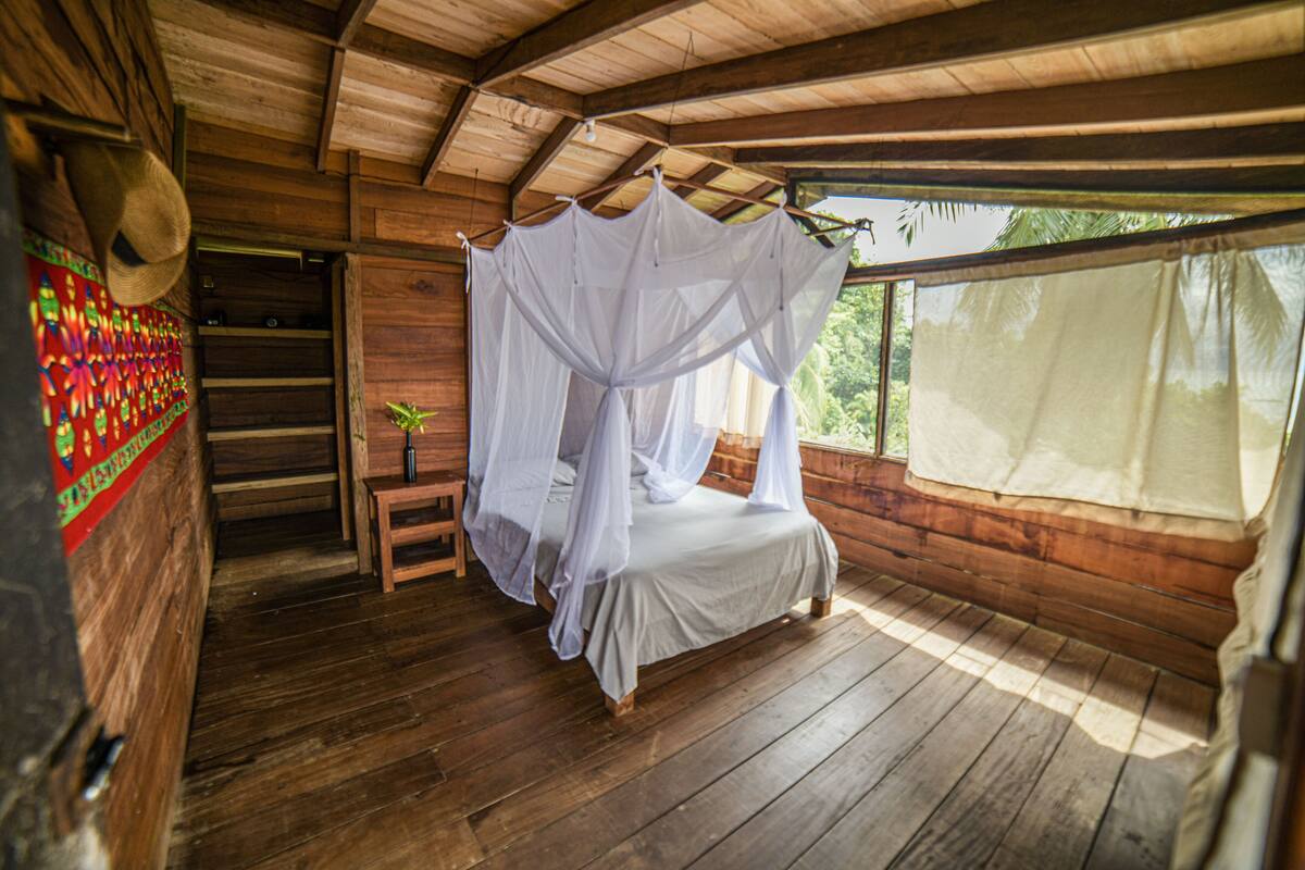 A wooden room with a bed under a white canopy, a small table with a plant, and a staircase leading to an upper level.