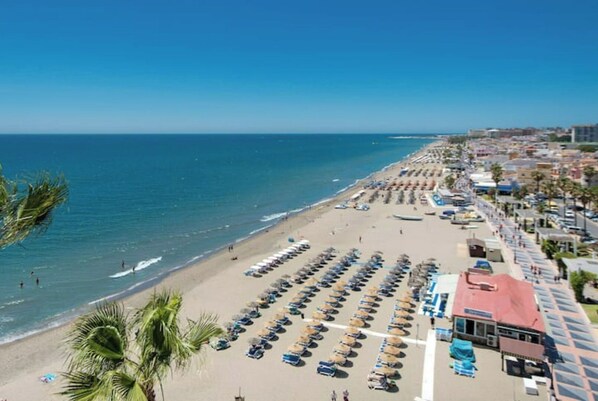 A beach with rows of umbrellas and sun loungers, a parking area, and a building with a red roof.