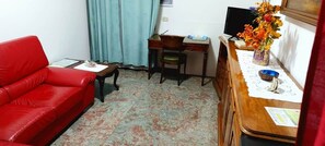 Ein Zimmer mit einem roten Sofa, einem kleinen Tisch, einem Schreibtisch mit Stuhl und einem Holzschrank.