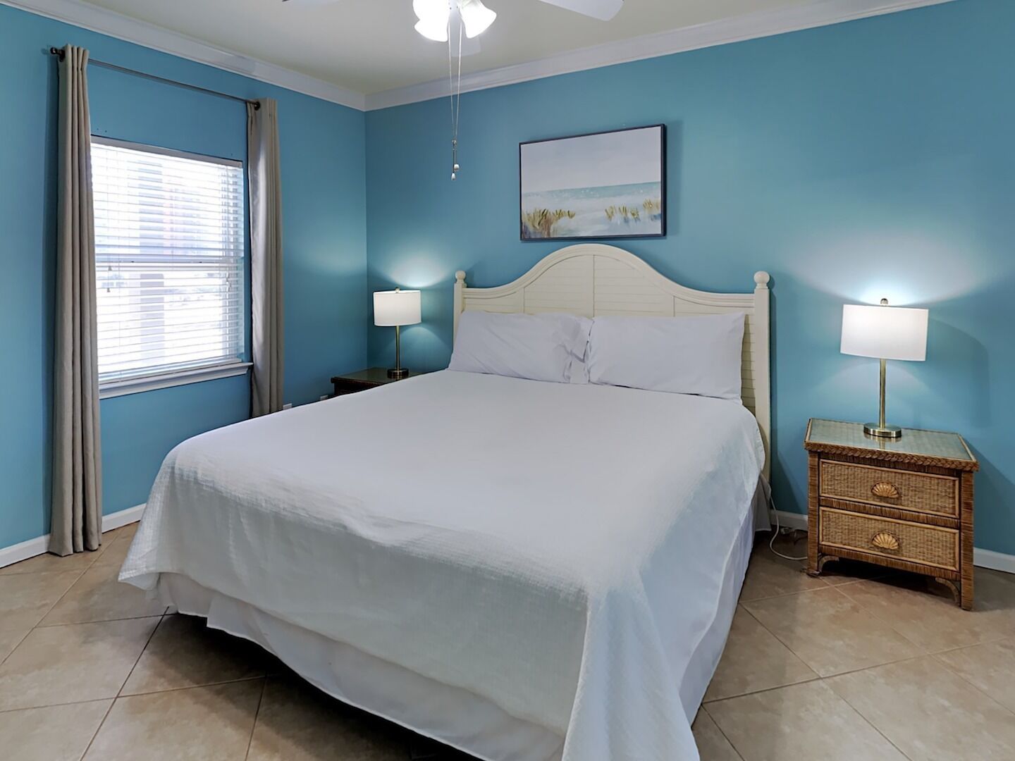 A bedroom with a large bed, two bedside tables with lamps, a ceiling fan, a window with curtains, and a painting on the wall.