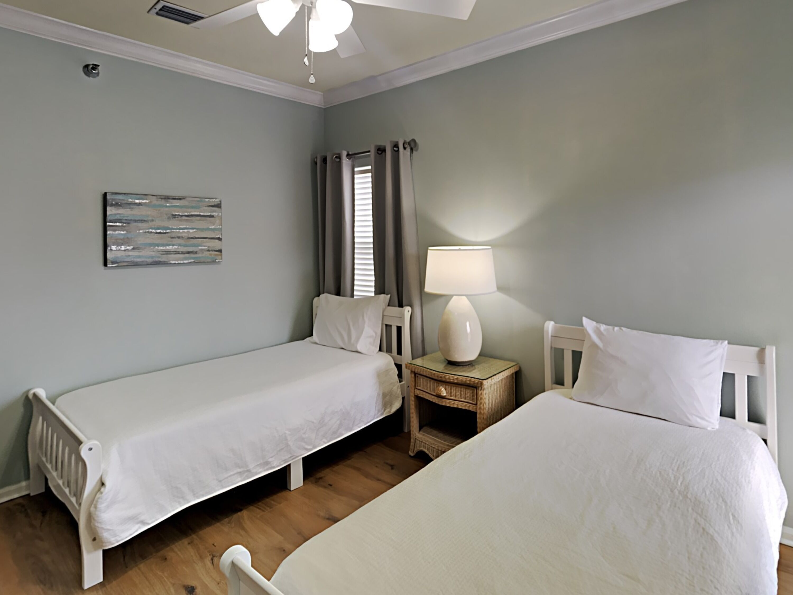 Une chambre avec deux lits simples, une table de chevet avec une lampe, un ventilateur de plafond et une œuvre d’art accrochée au mur.