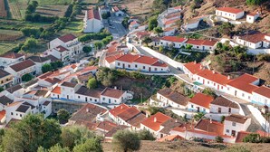 A hillside village with white buildings and red-tiled roofs, surrounded by green fields and trees.