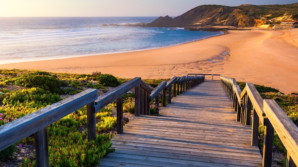 A wooden boardwalk leading to a beach with a clear view of the ocean and distant hills.