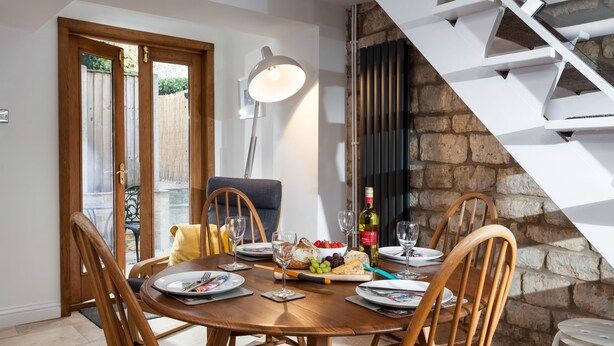 A dining area with a round wooden table set for four, surrounded by wooden chairs. A bottle of wine, a bowl of grapes, and a cheese board are on the table. A white lamp is on the wall, and a staircase with a stone wall is visible in the background. - Forge Cottage - Winchcombe, Cheltenham, a charming Cotswolds property sleeping 4 guests (Winchcombe)