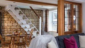 A cozy living room with a stone wall, a wooden staircase, a dining area with wooden chairs and a table, and a blue sofa with cushions. - Forge Cottage - Winchcombe, Cheltenham, a charming Cotswolds property sleeping 4 guests (Winchcombe)