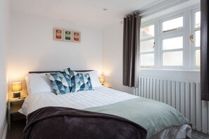 A neatly made bed with a white and green bedspread, two patterned pillows, and a dark throw blanket. There is a nightstand with a lamp and a framed certificate on the wall. - Forge Cottage - Winchcombe, Cheltenham, a charming Cotswolds property sleeping 4 guests (Winchcombe)
