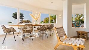 Een moderne eethoek met een grote tafel en rieten stoelen, grote ramen met uitzicht op zee, en een decoratieve vaas met pampasgras.