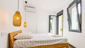 Een slaapkamer met een houten bed, witte beddengoed, twee hanglampen, een airconditioner en een glazen schuifdeur naar een tuin.