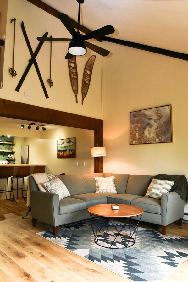 Un salon avec un canapé d’angle gris, une table basse ronde, un ventilateur de plafond et un tableau accroché au mur.
