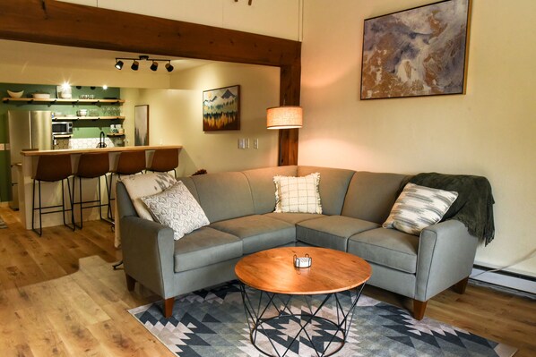 A living room with a gray sectional sofa, a wooden coffee table, and a kitchen in the background.