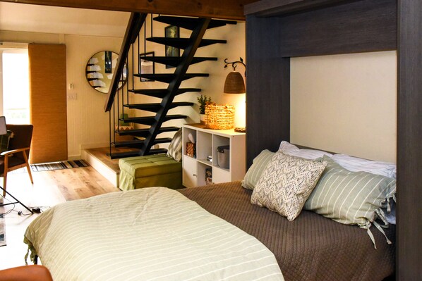 A bedroom with a bed, a chair, a staircase, and a shelf with decorative items.