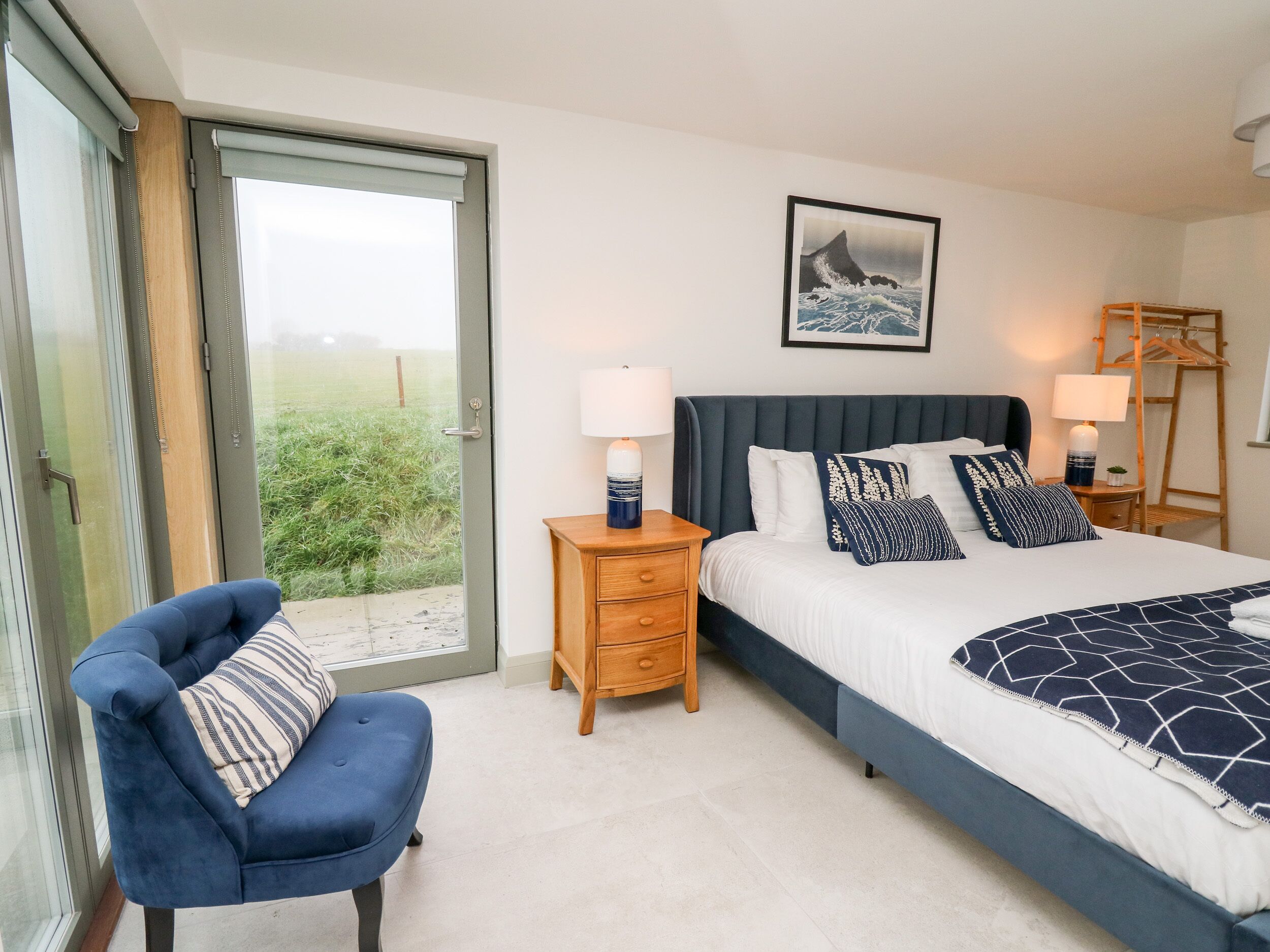A bedroom with a bed, a chair, a nightstand, a lamp, a framed picture, and a view through a glass door.