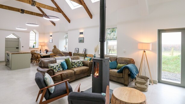 A spacious living room with a high ceiling, exposed wooden beams, a central fireplace, and a view of the outdoors through large windows. - Ysgoldy Brynwyre (Llanon)