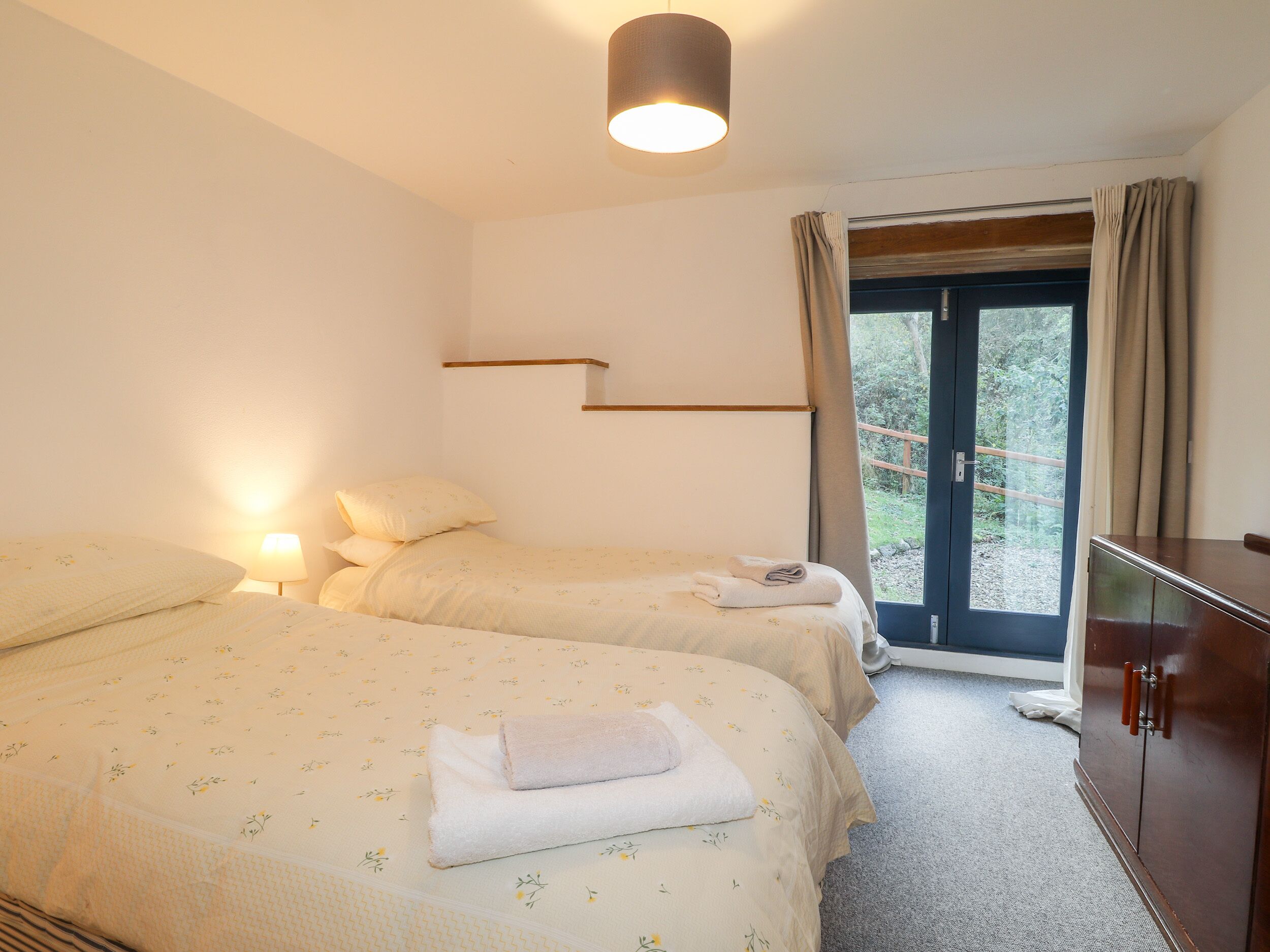 Quarto com duas camas, luminária de cabeceira, guarda-roupa e janela com cortinas.
