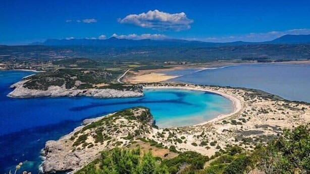 Paisaje costero con una playa, aguas azules cristalinas y montañas lejanas.