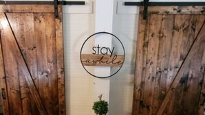A room with wooden sliding doors, a small black table with a potted plant, and a circular sign with the words "stay awhile". - Clemson Memorabilia House (Clemson)