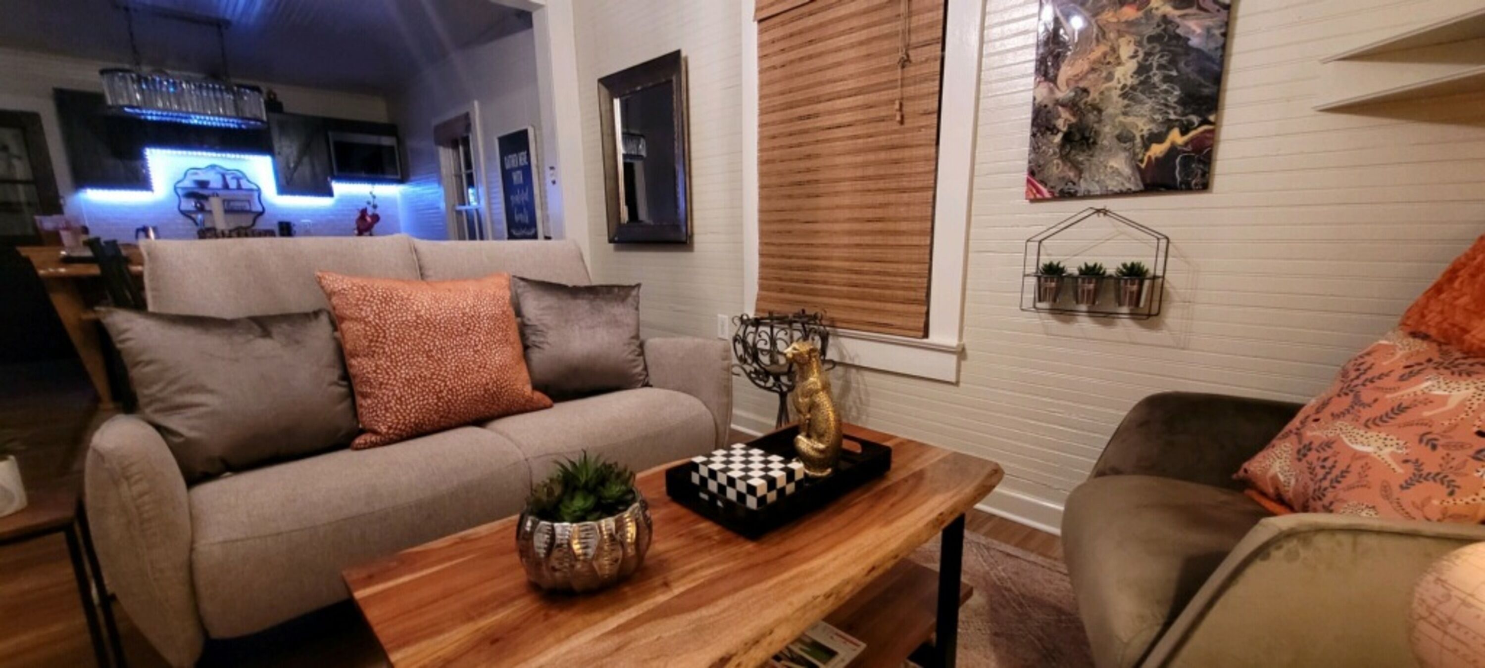 A living room with a sofa, a wooden coffee table, and a decorative chess piece. There is a window with blinds, a wall-mounted shelf with plants, and a painting.