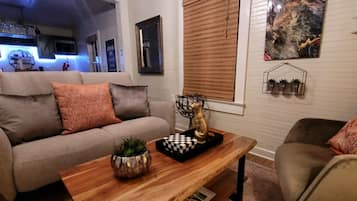 A living room with a sofa, a wooden coffee table, and a decorative chess piece. There is a window with blinds, a wall-mounted shelf with plants, and a painting.