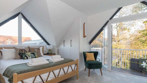 Un dormitorio con techo inclinado, una cama con estructura de madera, una silla verde y un balcón con vistas a árboles y edificios.