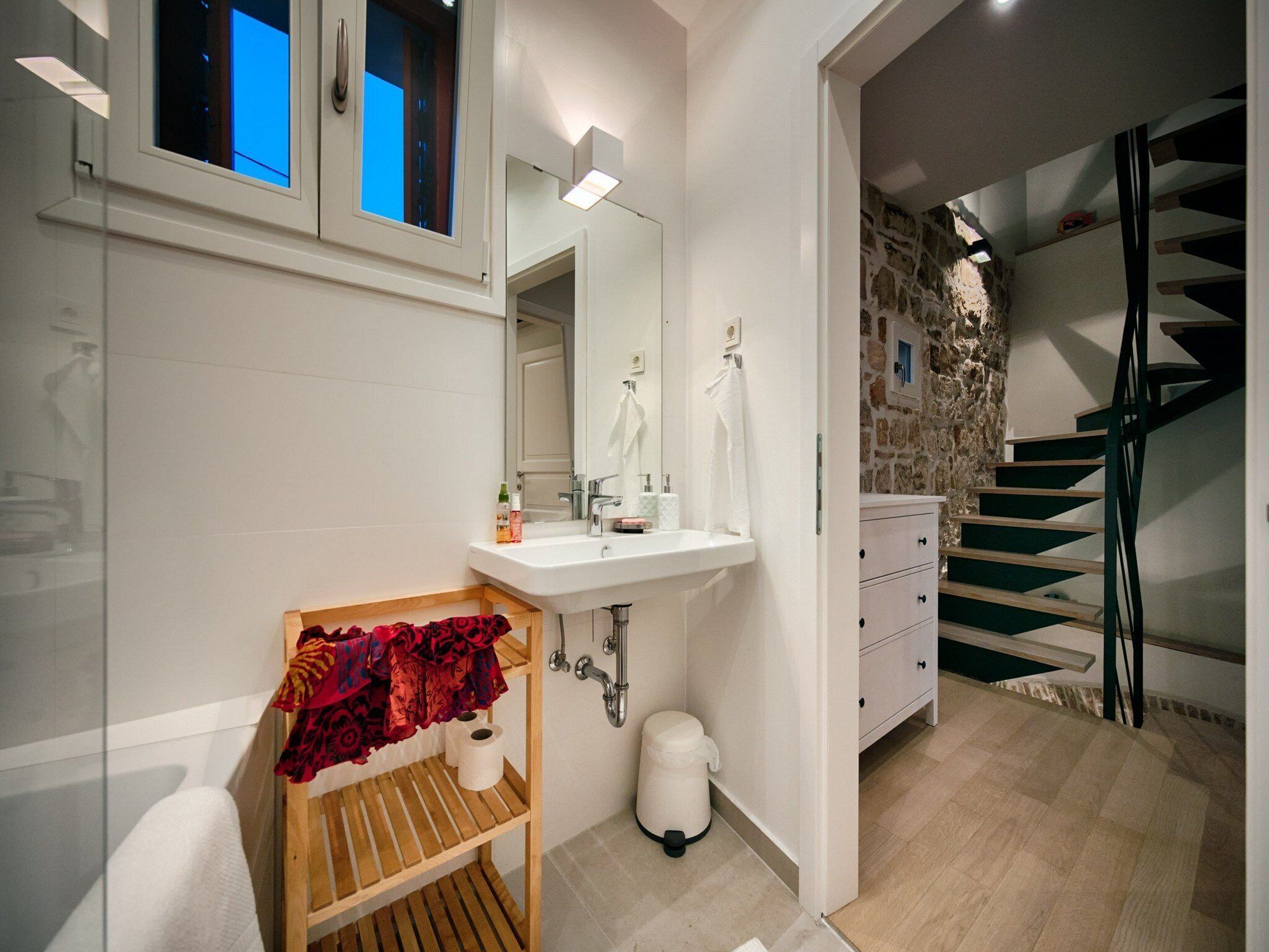 A modern bathroom with a white sink, mirror, and wooden shelf with towels. A stone wall and staircase are visible in the background.