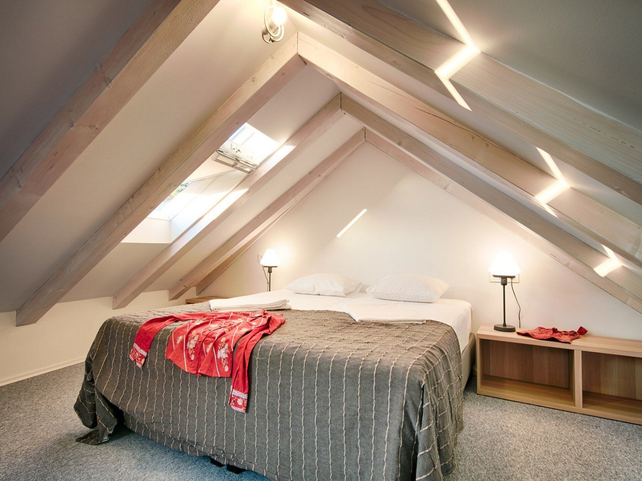 A bedroom with a sloped ceiling, a bed with a grey striped bedspread, two bedside lamps, and a small wooden nightstand.