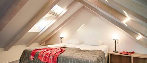 A bedroom with a sloped ceiling, a bed with a grey striped bedspread, two bedside lamps, and a small wooden nightstand.