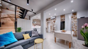 Un espace de vie moderne comprenant un canapé gris, une petite table ronde, un coin repas avec une table et des chaises blanches, et une cuisine avec un mur en briques.