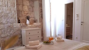 Un dormitorio con pared de piedra, una mesita de noche con un jarrón, una bandeja con fruta y una bebida, y una cama hecha con esmero.