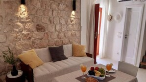Una habitación acogedora con pared de piedra, un sofá con cojines, una mesa de comedor con frutas y bebidas, y un florero con plantas.