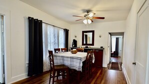 Comedor con mesa lista para cuatro, ventilador de techo y zona de cocina con espejo y armarios.