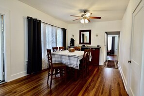 Comedor con mesa lista para cuatro, ventilador de techo y zona de cocina con espejo y armarios.