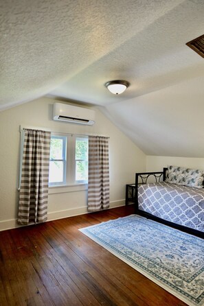 A bedroom with a sloped ceiling, an air conditioning unit, a window with curtains, a bed with patterned bedding, and a blue patterned rug.
