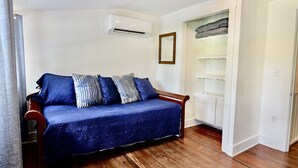 Una habitación con suelo de madera, un sofá azul con cojines estampados, un equipo de aire acondicionado blanco y una puerta blanca con un espejo.
