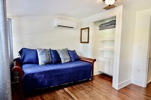 Una habitación con suelo de madera, un sofá azul con cojines estampados, un equipo de aire acondicionado blanco y una puerta blanca con un espejo.