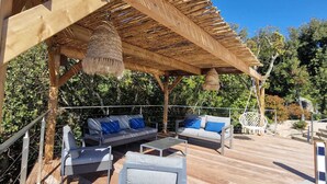Una terraza de madera con mobiliario exterior, incluyendo sofás y una mesa, bajo una pérgola con lámparas colgantes.