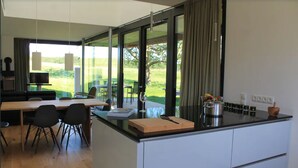 A modern kitchen with a large island, dining area with chairs, and large windows offering a view of the outdoors. - Thatched roof meets modernity; lake view, sauna, garden and much more... (Putbus)