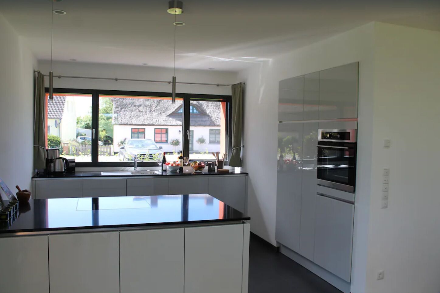 Une cuisine moderne dotée d’un grand îlot central, d’un four encastré et offrant une vue sur une maison par la fenêtre.
