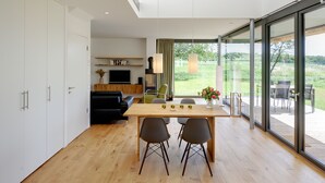 A modern kitchen with a dining area, wooden flooring, and a large sliding glass door leading to an outdoor patio. - Thatched roof meets modernity; lake view, sauna, garden and much more... (Putbus)