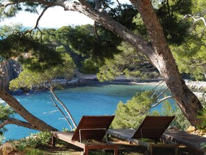 A secluded beach area with two wooden lounge chairs facing a calm sea, surrounded by dense pine trees and rocky terrain. - Waterfront, sleeps 8, direct access to the sea in 1 min.  (Ensuès-la-Redonne)