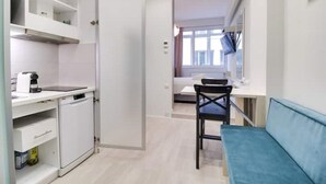 Een compacte kitchenette met een koffiezetapparaat, een kleine koelkast en een gootsteen. In de woonkamer staat een blauwe bank en een stoel, met een raam dat voor natuurlijk licht zorgt.