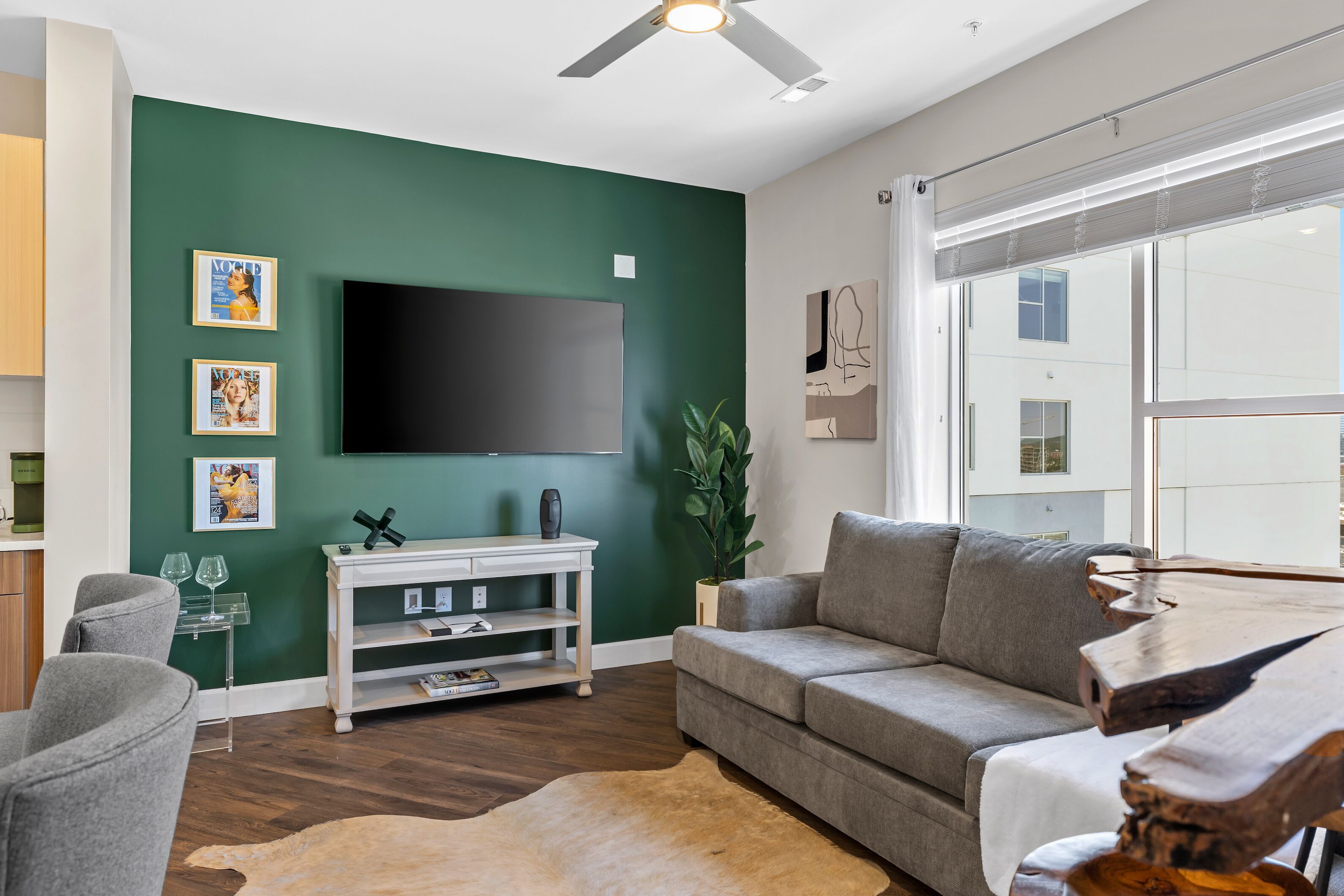 A modern living room with a green accent wall, a flat-screen TV, a grey sofa, a wooden coffee table, and a large window.
