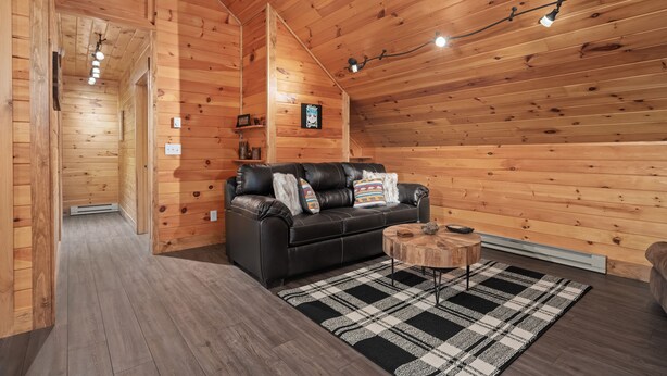 A cozy wooden cabin interior with a black leather sofa, a plaid rug, and a wooden coffee table. - Loft In the Pines, On Snowmobile Trail (Boonville)