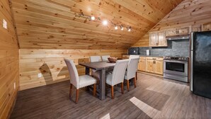 A wooden interior with a dining table and chairs, a kitchen with wooden cabinets and appliances, and a sloped ceiling. - Loft In the Pines, On Snowmobile Trail (Boonville)