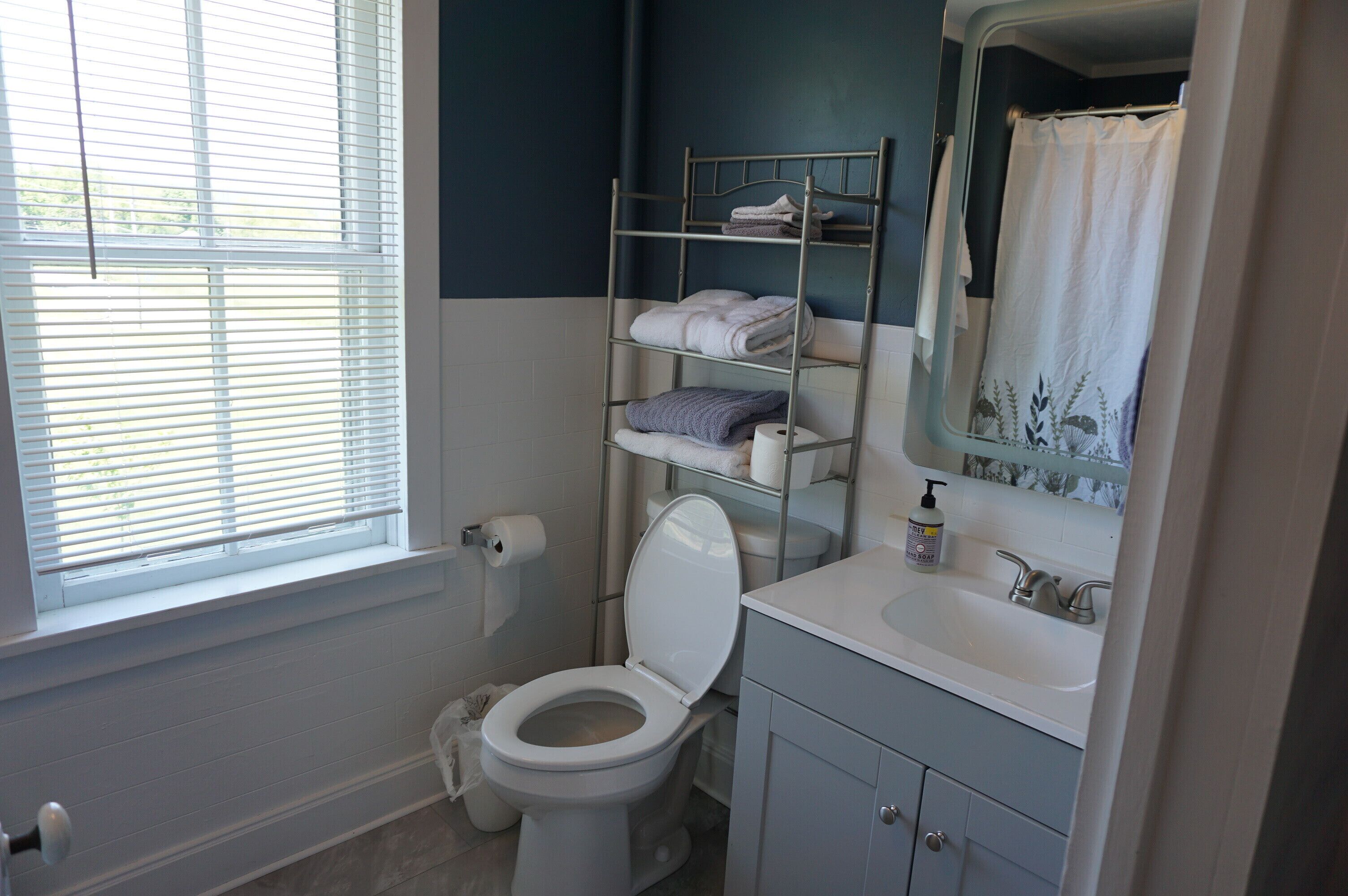 A bathroom with a toilet, a sink, a mirror, and a window with blinds.