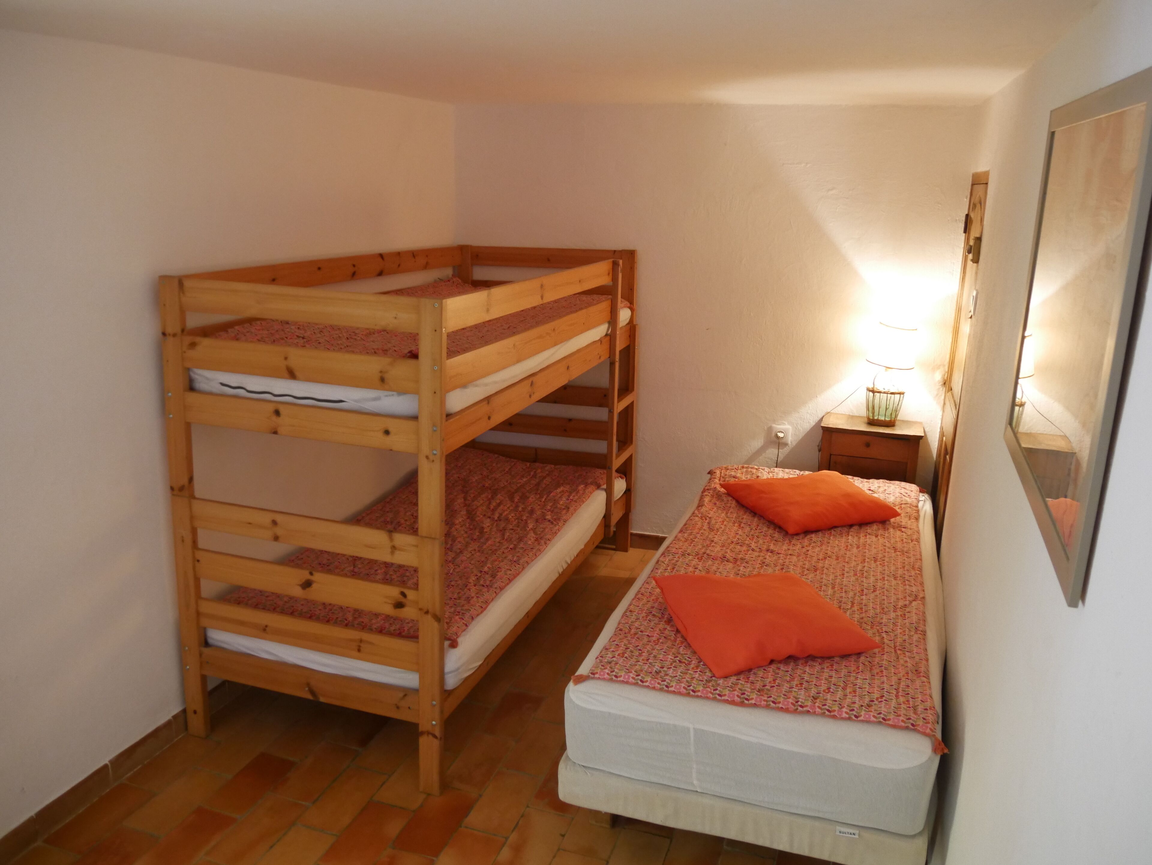 A room with a bunk bed, a single bed, a nightstand with a lamp, and a mirror on the wall.