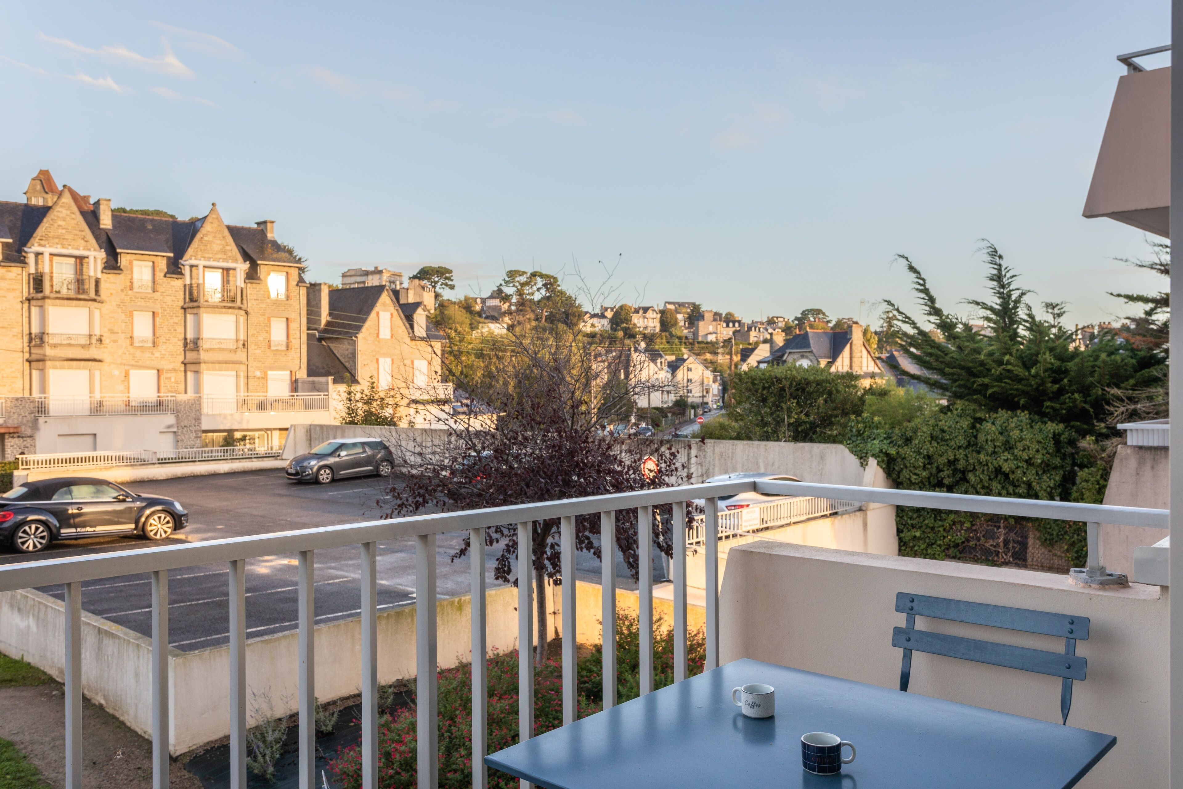 Un balcon avec une table et deux tasses, donnant sur une rue bordée de voitures garées et d’immeubles d’habitation.