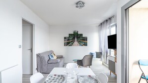 A modern dining area with a table set for two, a grey sofa, a TV, and a painting of a bridge. - Marée Tropicale - vue mer (Saint-Cast-le-Guildo)