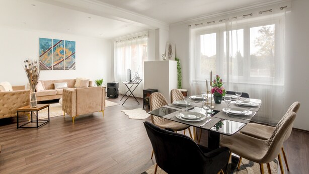 A modern living room with a dining area, featuring a sofa set, a glass-top dining table, and a large window with curtains.